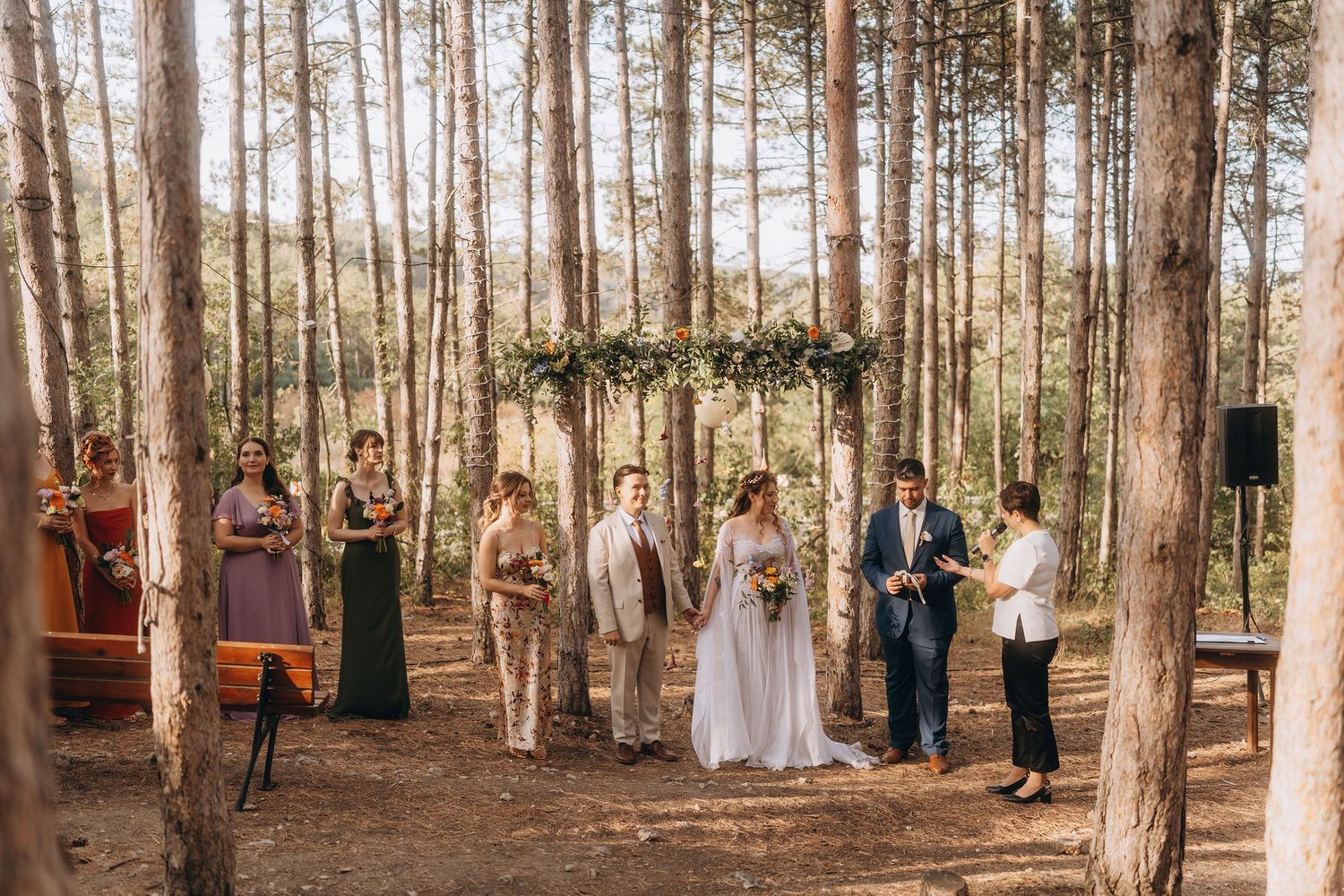 Outdoor forest wedding ceremony with bridal party standing among tall pine trees at a rustic woodland altar.