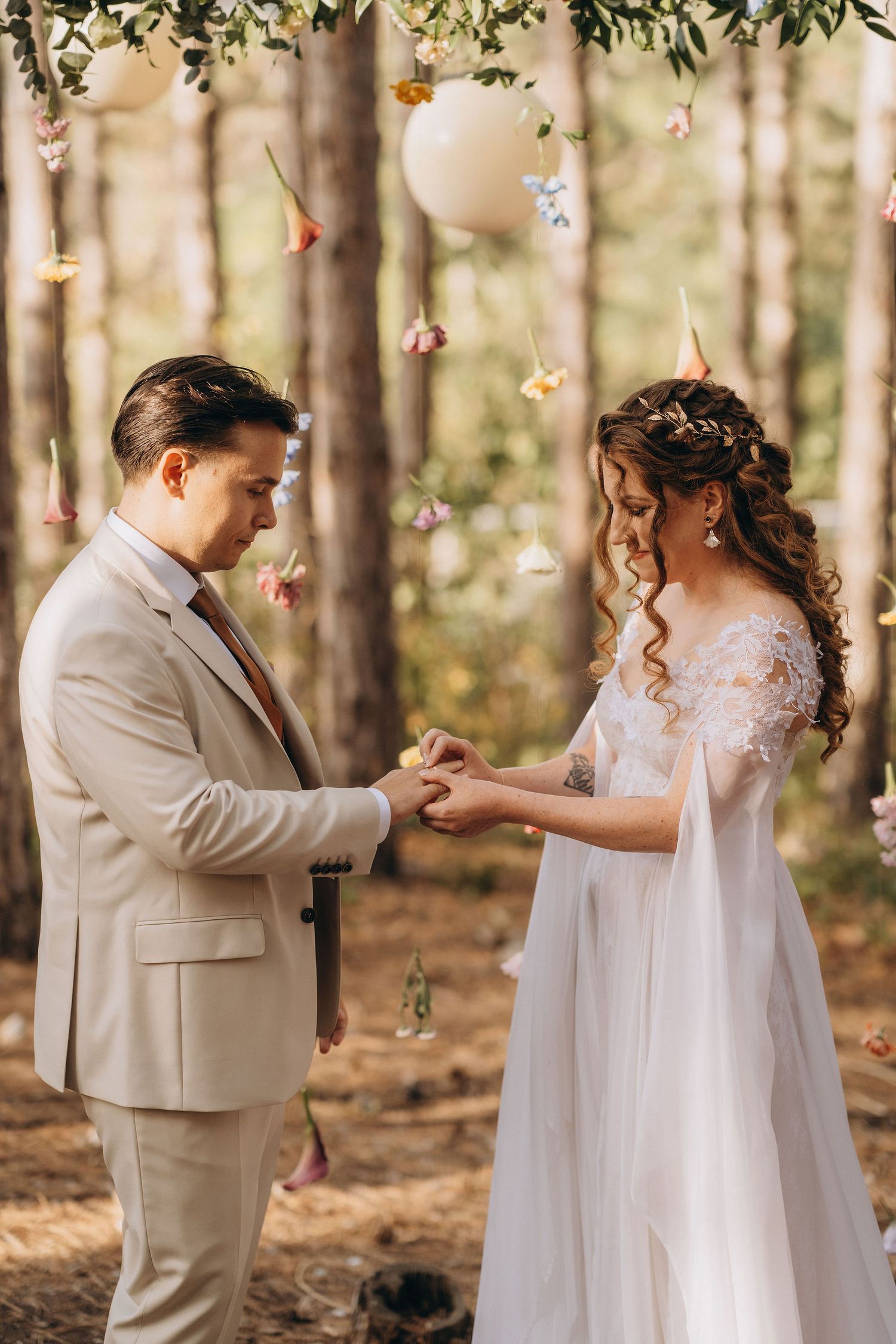 Groom placing wedding ring on bride's finger during outdoor forest ceremony with floral decorations.