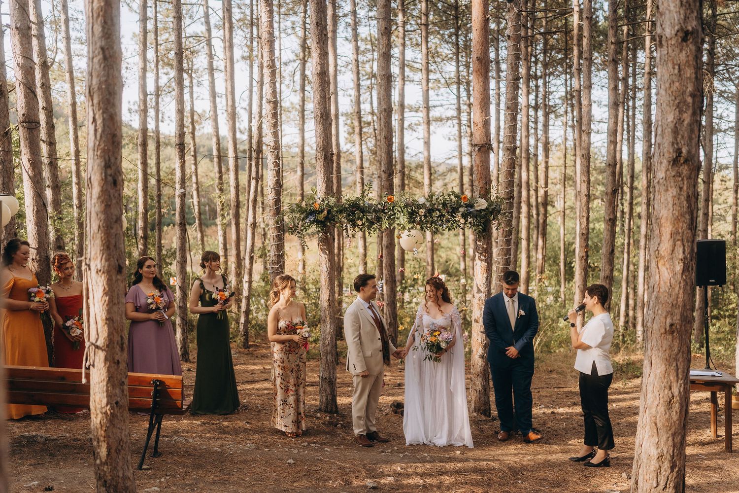 Outdoor forest wedding ceremony with bride and groom at floral arch surrounded by bridesmaids and officiant among tall pine trees.