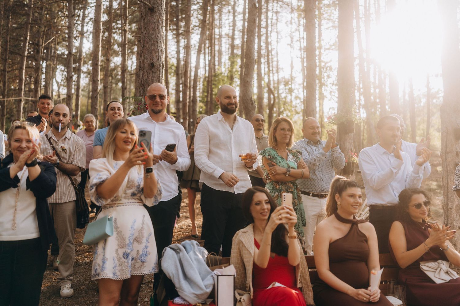 Wedding guests gathered in sunlit forest clearing, applauding and smiling during outdoor ceremony celebration.