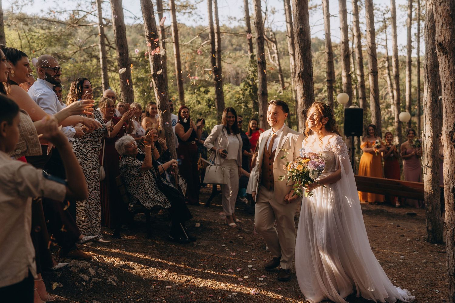 Newlywed couple walks down forest aisle hand-in-hand as guests celebrate their outdoor woodland wedding ceremony.