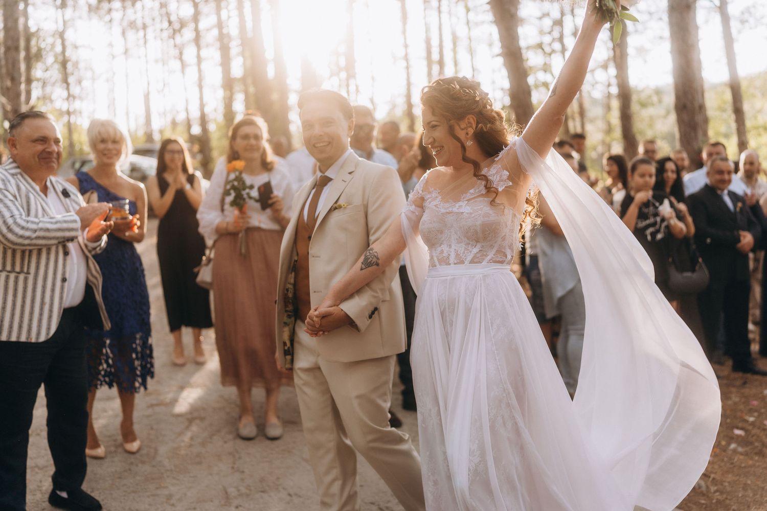 Joyful bride raises arm in celebration while walking with groom through forest wedding surrounded by applauding guests.