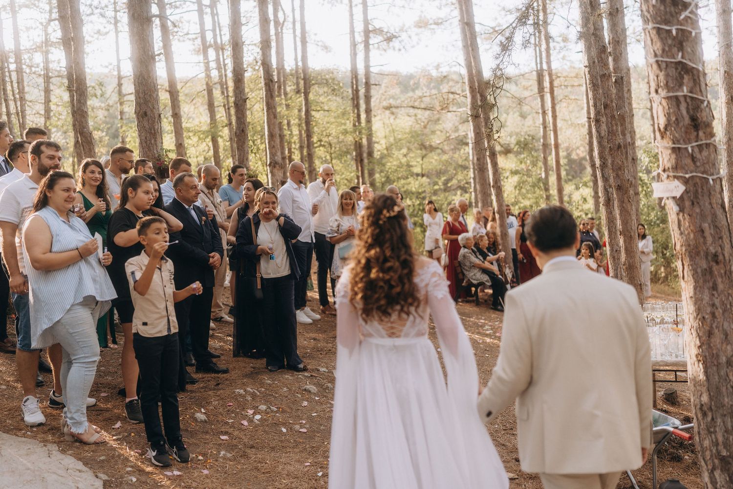 Bride and groom face their guests during an outdoor forest wedding celebration among tall pine trees in natural sunlight.