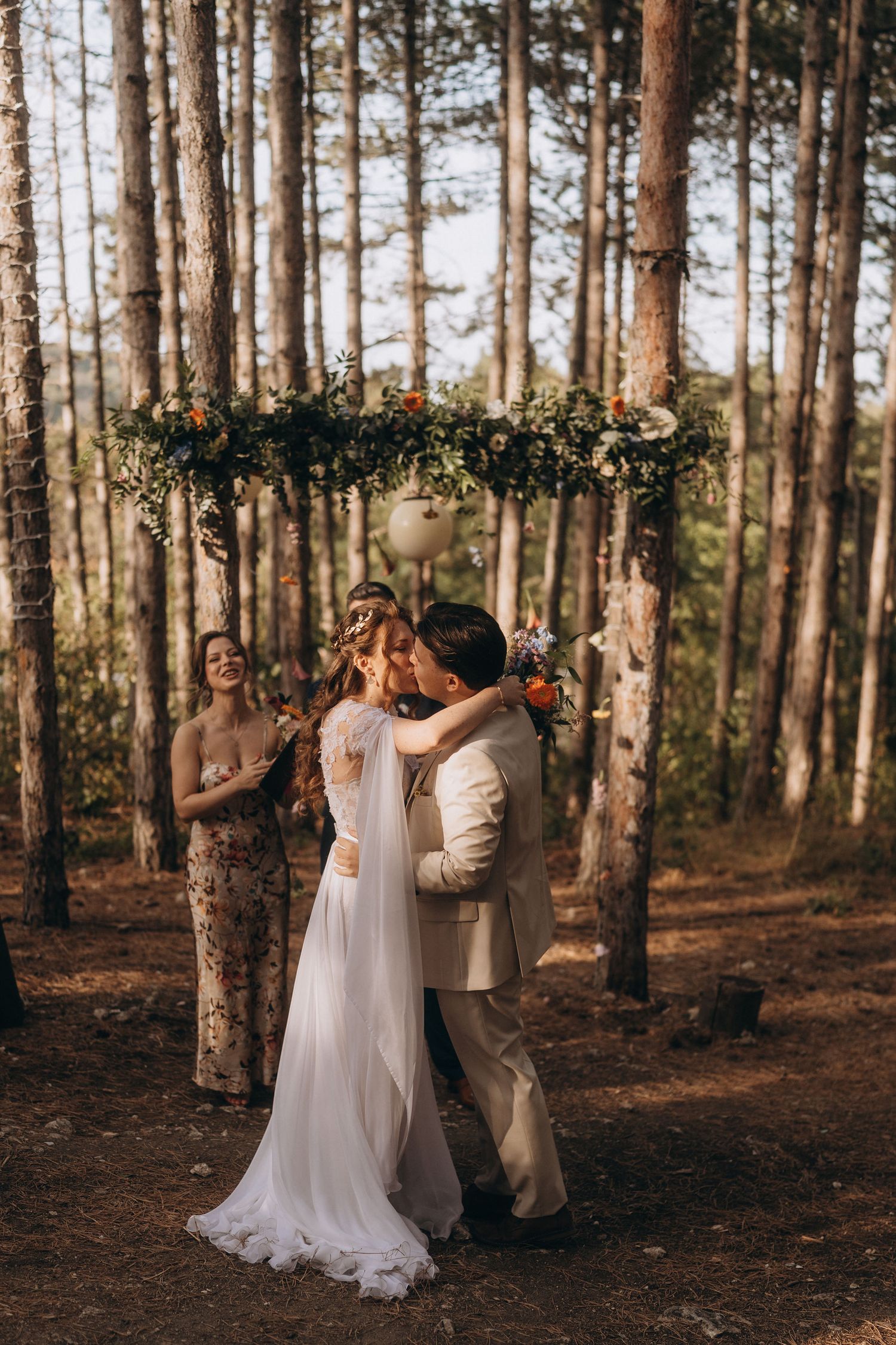 Bride and groom share first kiss under floral arch during romantic outdoor wedding ceremony in pine forest.