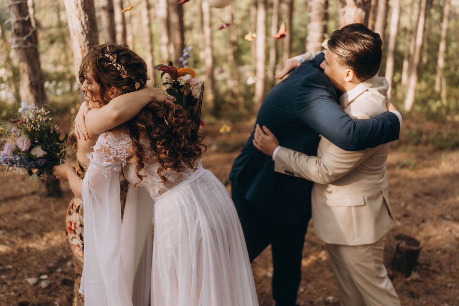 Wedding party exchanges warm hugs after ceremony in sunlit pine forest, bride in white gown and groom in beige suit.