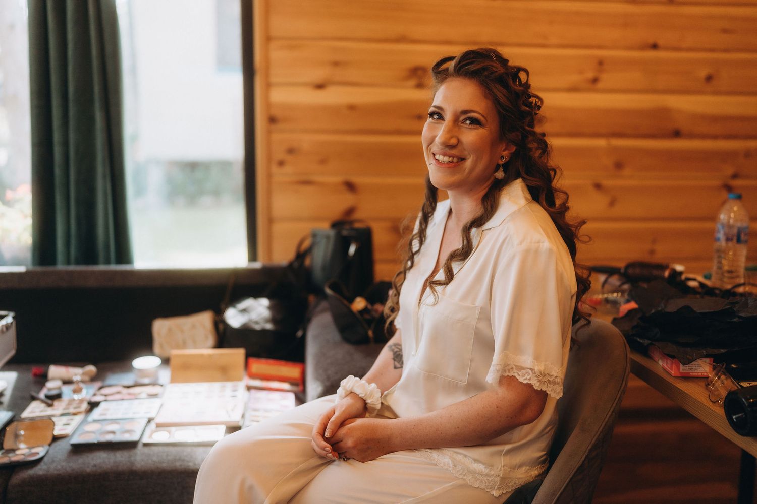 Bride in white robe sitting at vanity table smiling, getting ready in a rustic wooden cabin.
