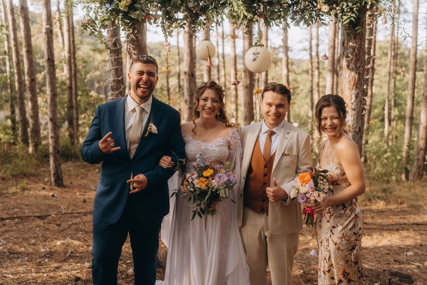 Two couples pose joyfully together in a forest wedding ceremony setting with balloon decorations.