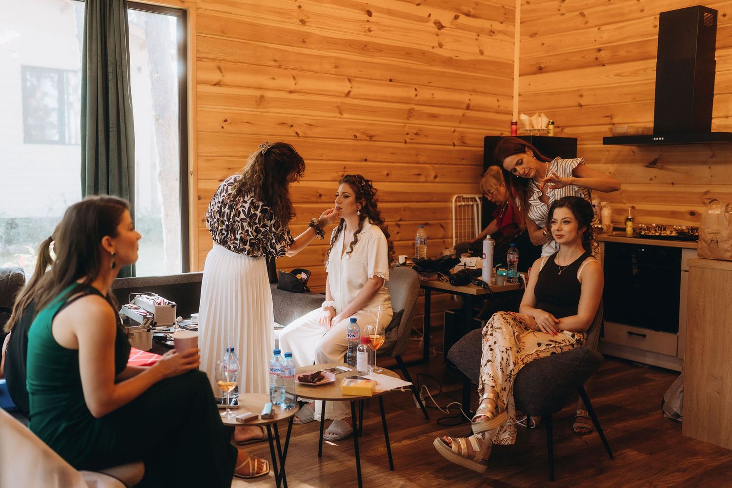 Women gather in a cozy wooden cabin for bridal party preparations, with makeup and hair styling in progress.