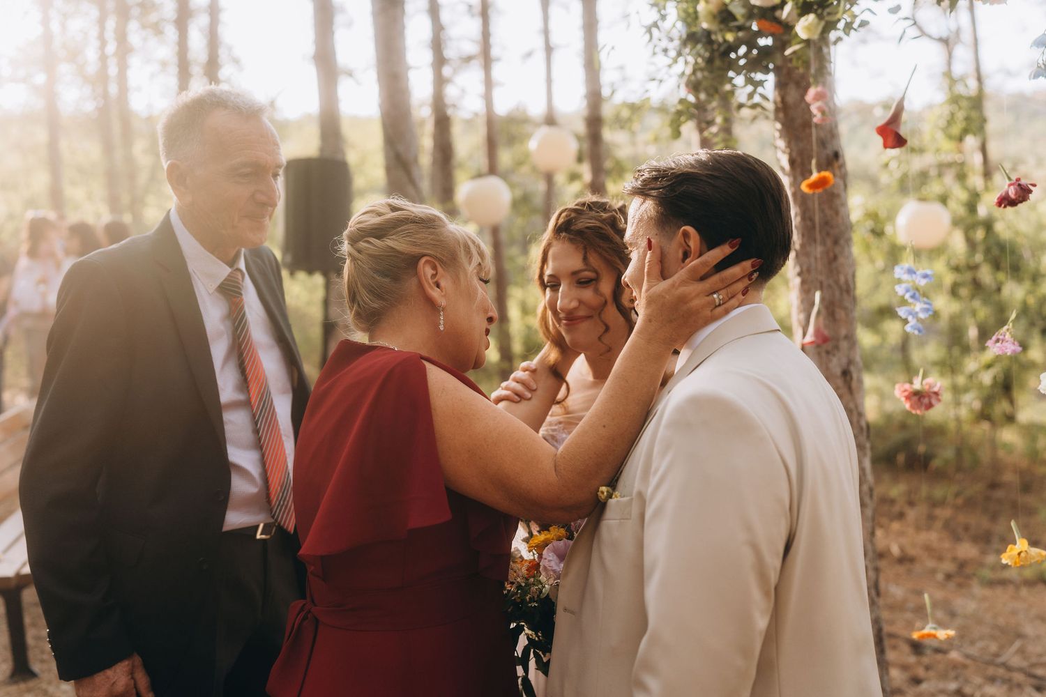 Emotional mother in red dress tenderly cups grooms face while bride looks on during outdoor forest wedding celebration.