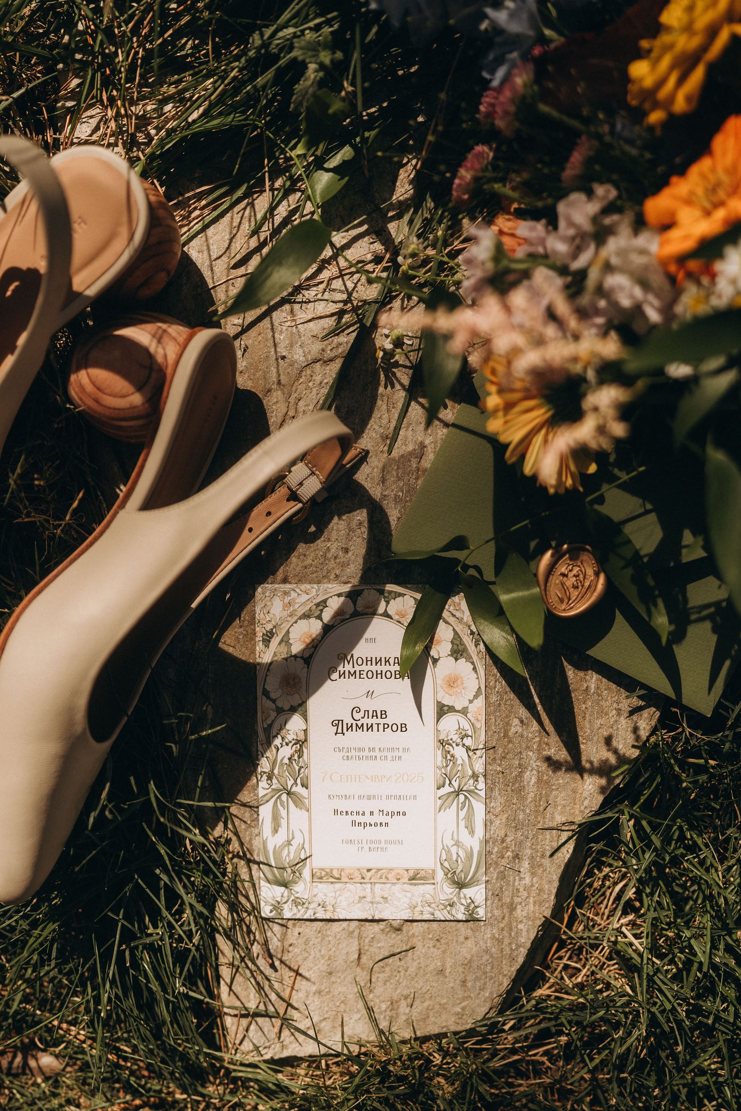 Wedding flat lay with invitation cards, nude heels, wax seal, and colorful floral bouquet on grass and stone.