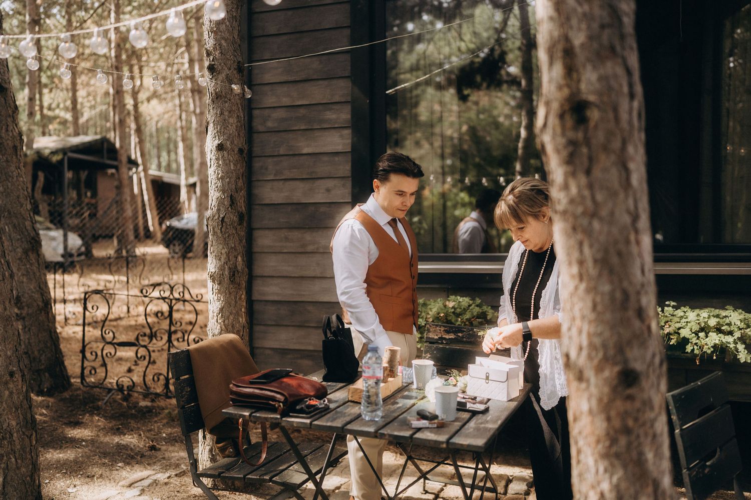 A man in a vest and woman in black set an outdoor cafe table surrounded by pine trees and string lights.