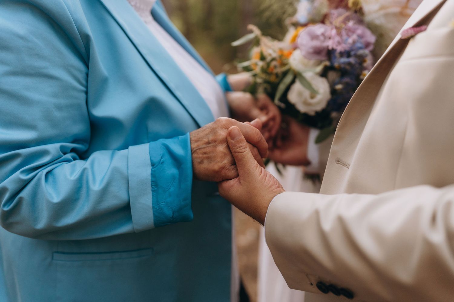 Close-up of two people exchanging rings at wedding ceremony, one wearing blue suit holding colorful bouquet.