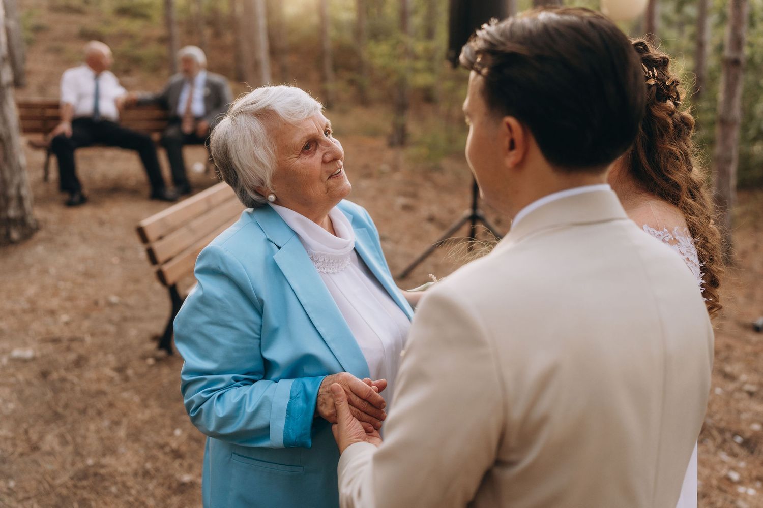 Elderly woman in blue jacket holds hands with groom during outdoor woodland wedding ceremony.