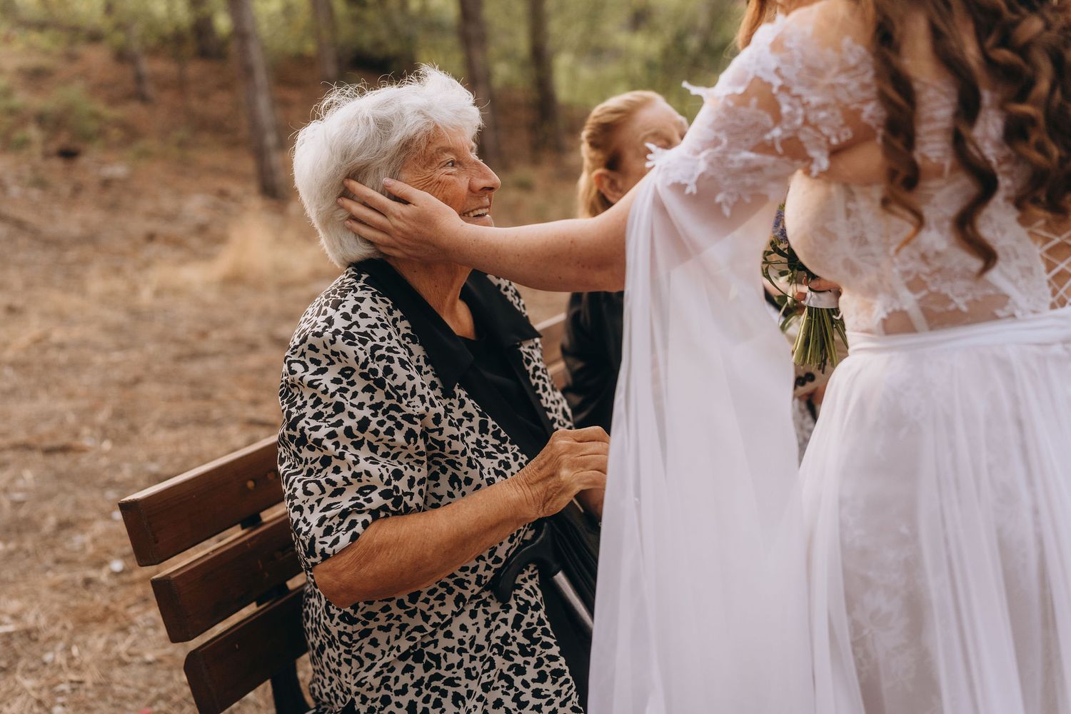An elderly woman seated on a bench tenderly touches a bride's face in an outdoor forest setting.