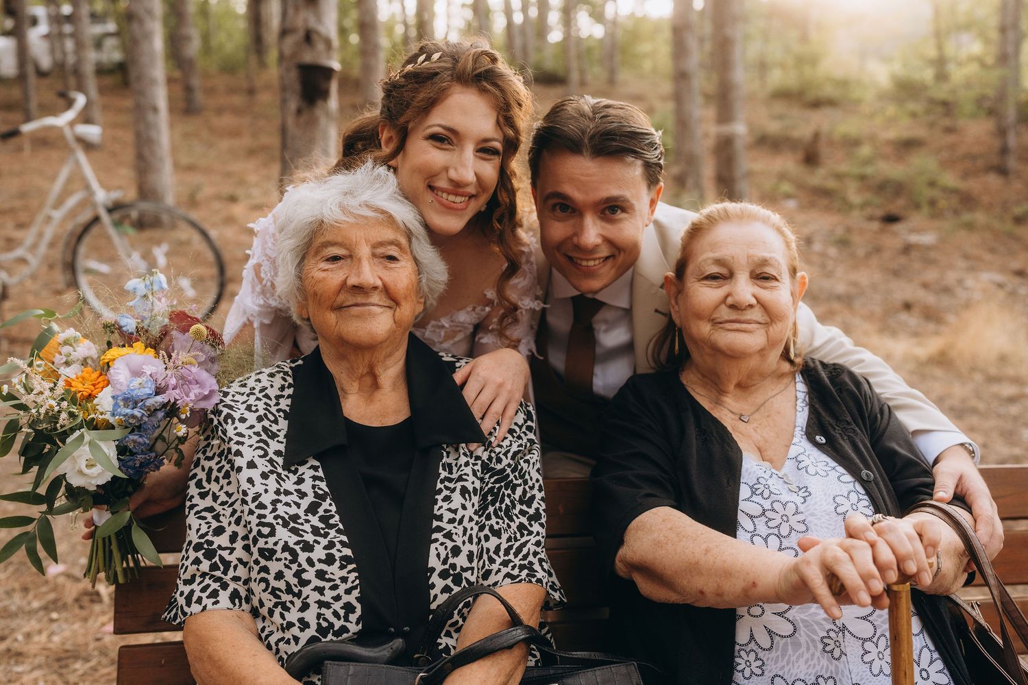 Wedding couple poses with two elderly grandmothers in a forest setting holding bouquet.