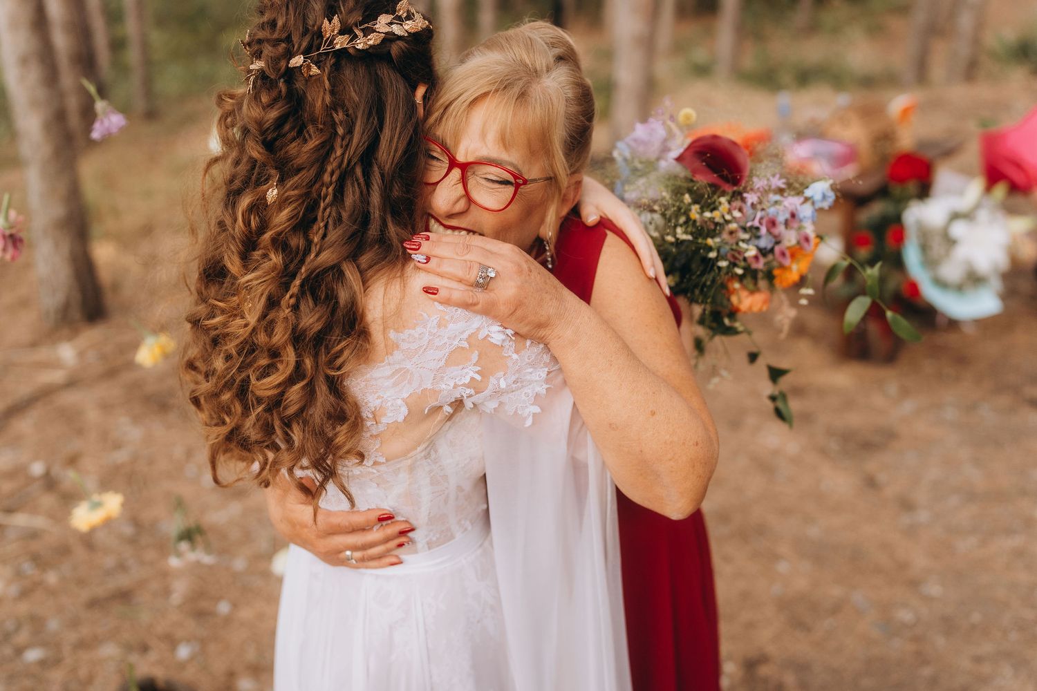 Older woman in red dress embracing bride in white lace gown at outdoor forest wedding.