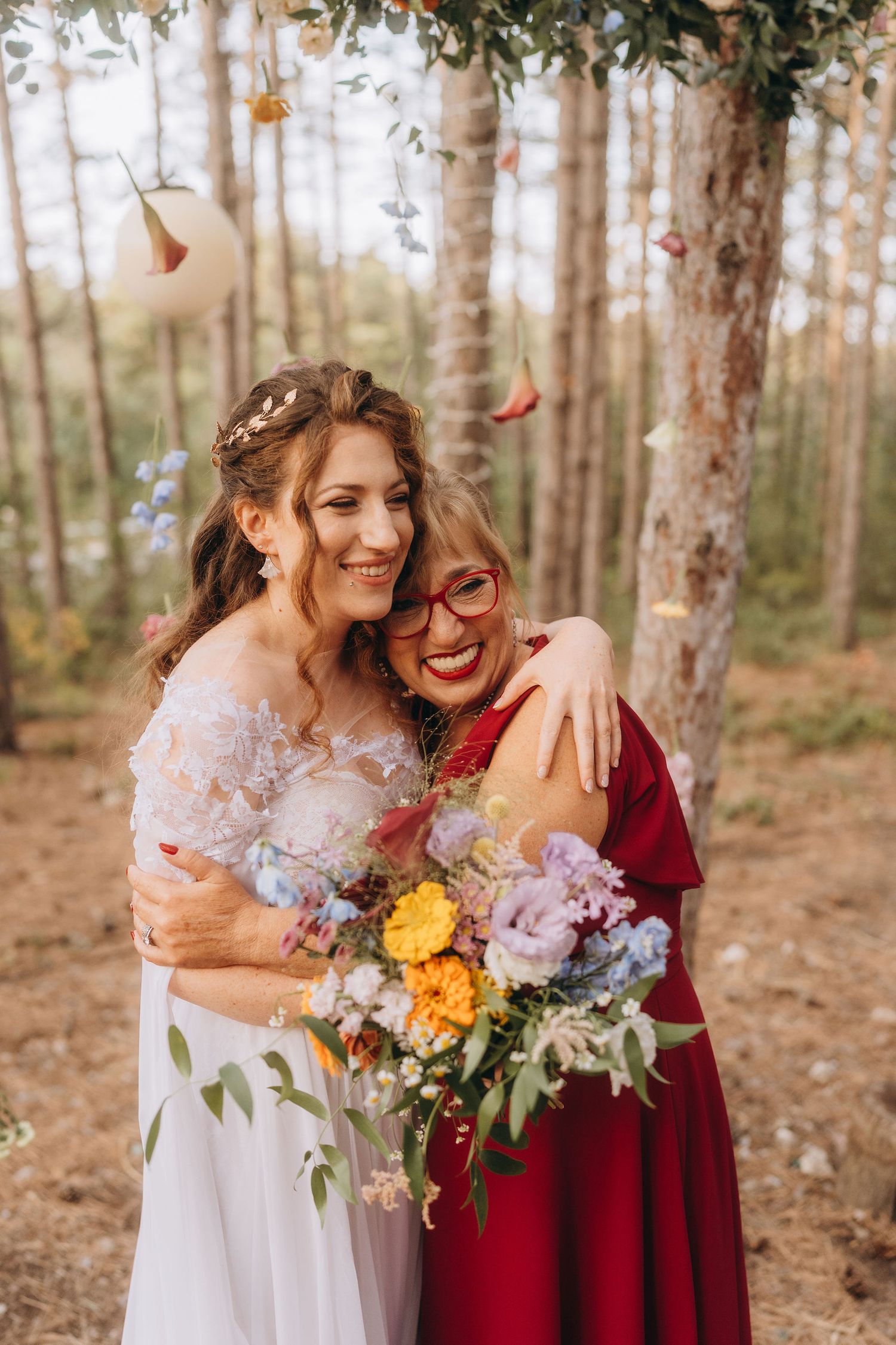 A bride in a white lace gown hugs a woman in a red dress while holding a colorful bouquet outdoors.