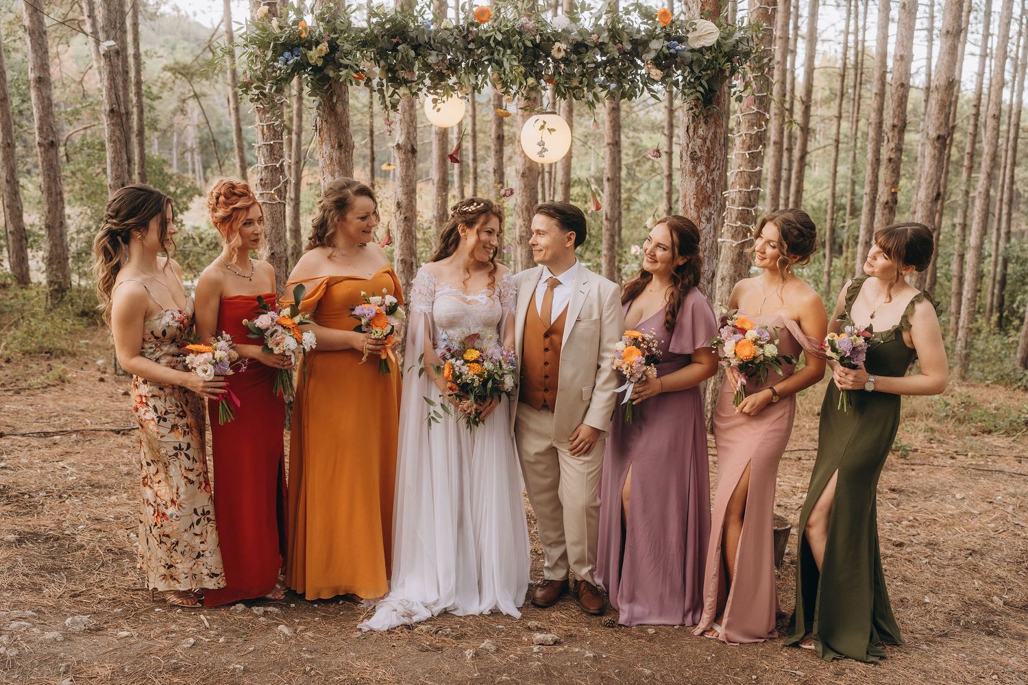Wedding party with bride, groom, and six bridesmaids in colorful dresses posing in forest setting.