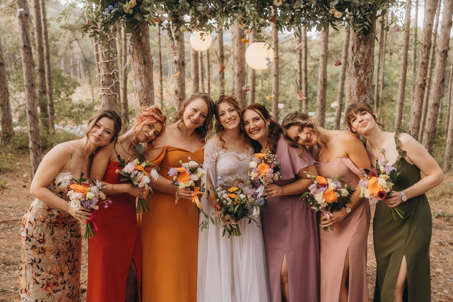 Seven bridesmaids in colorful mismatched dresses holding bouquets at a forest wedding.