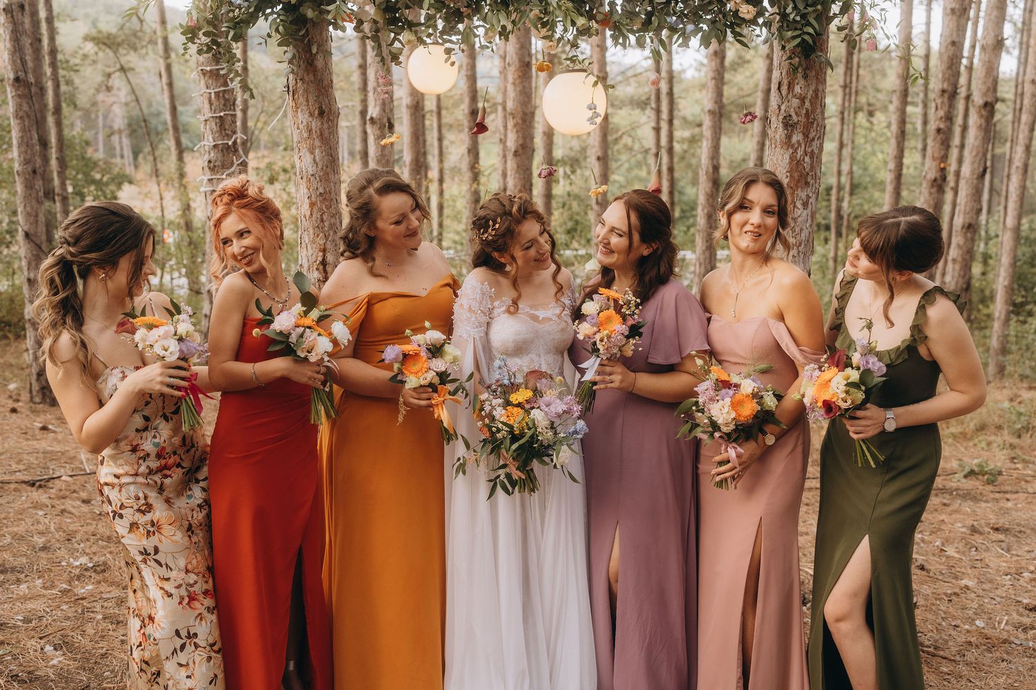 A bride in white stands with six bridesmaids in colorful mismatched dresses holding floral bouquets.