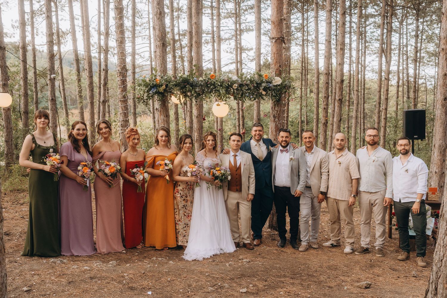 Wedding party poses together in a pine forest with floral arch, bridesmaids in earthy toned gowns.