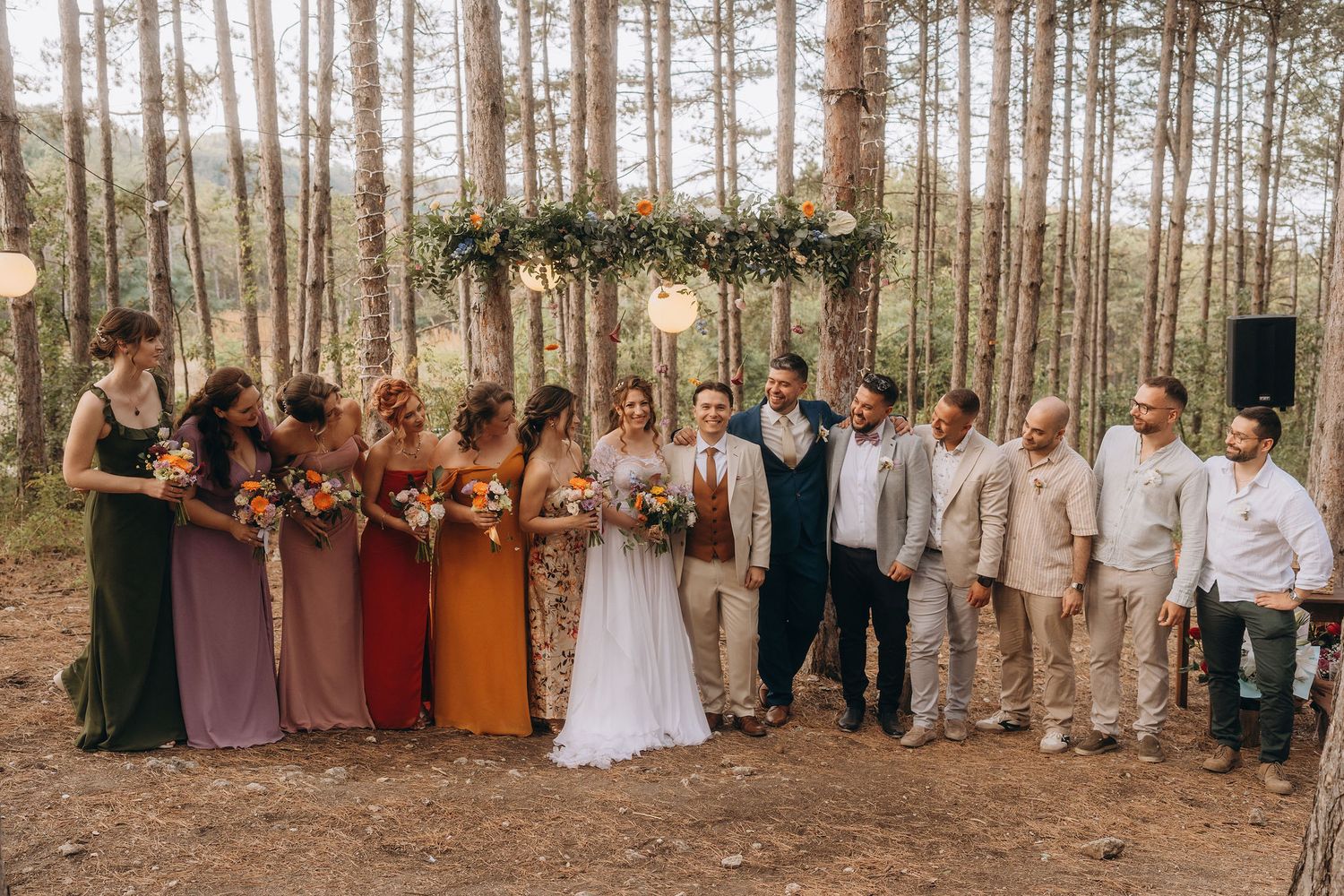 Large wedding party with bridesmaids in colorful gowns and groomsmen posing in pine forest.