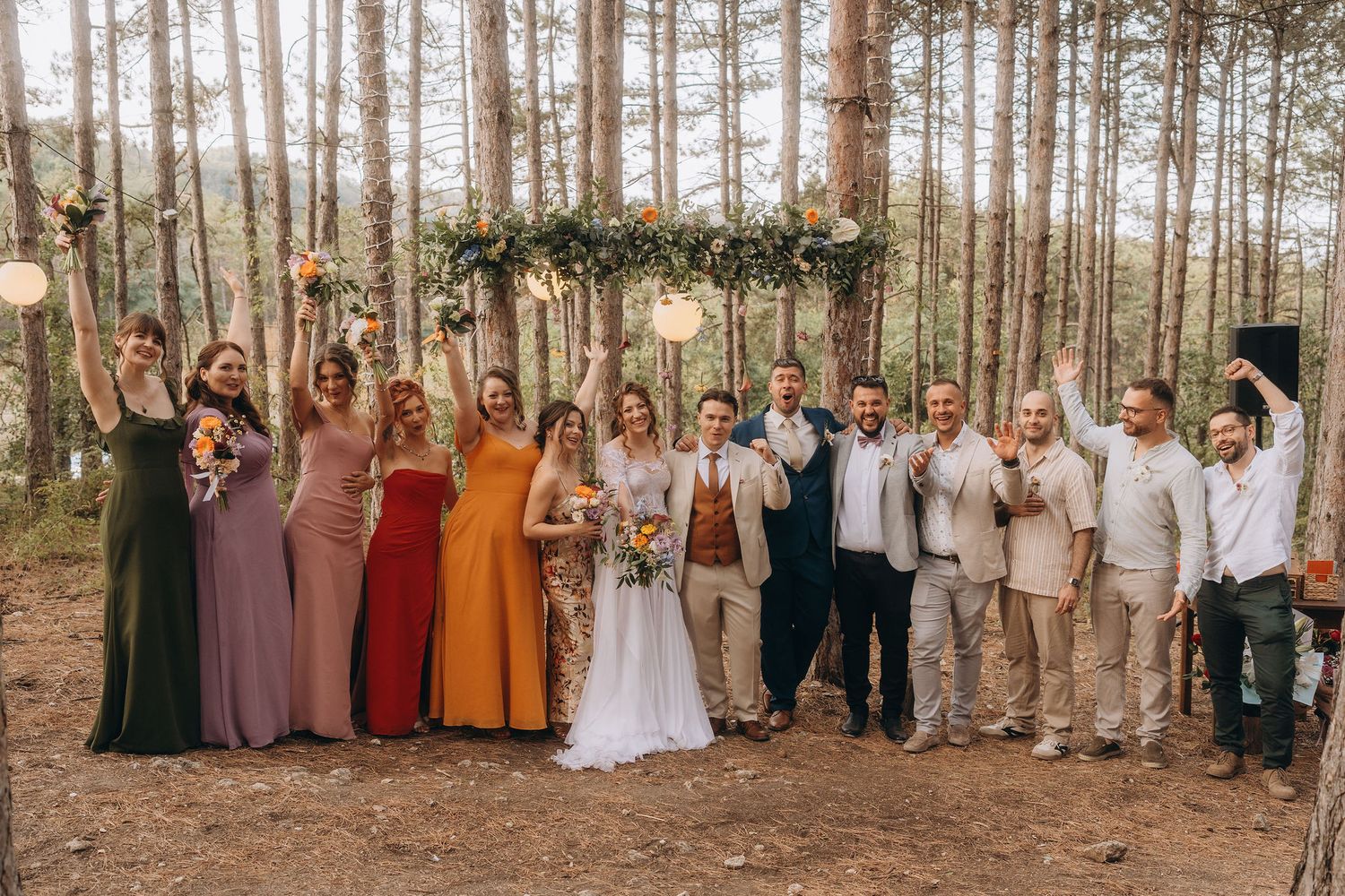 Wedding party celebrating in a pine forest with bridesmaids in earthy tones and groomsmen under a floral arch.