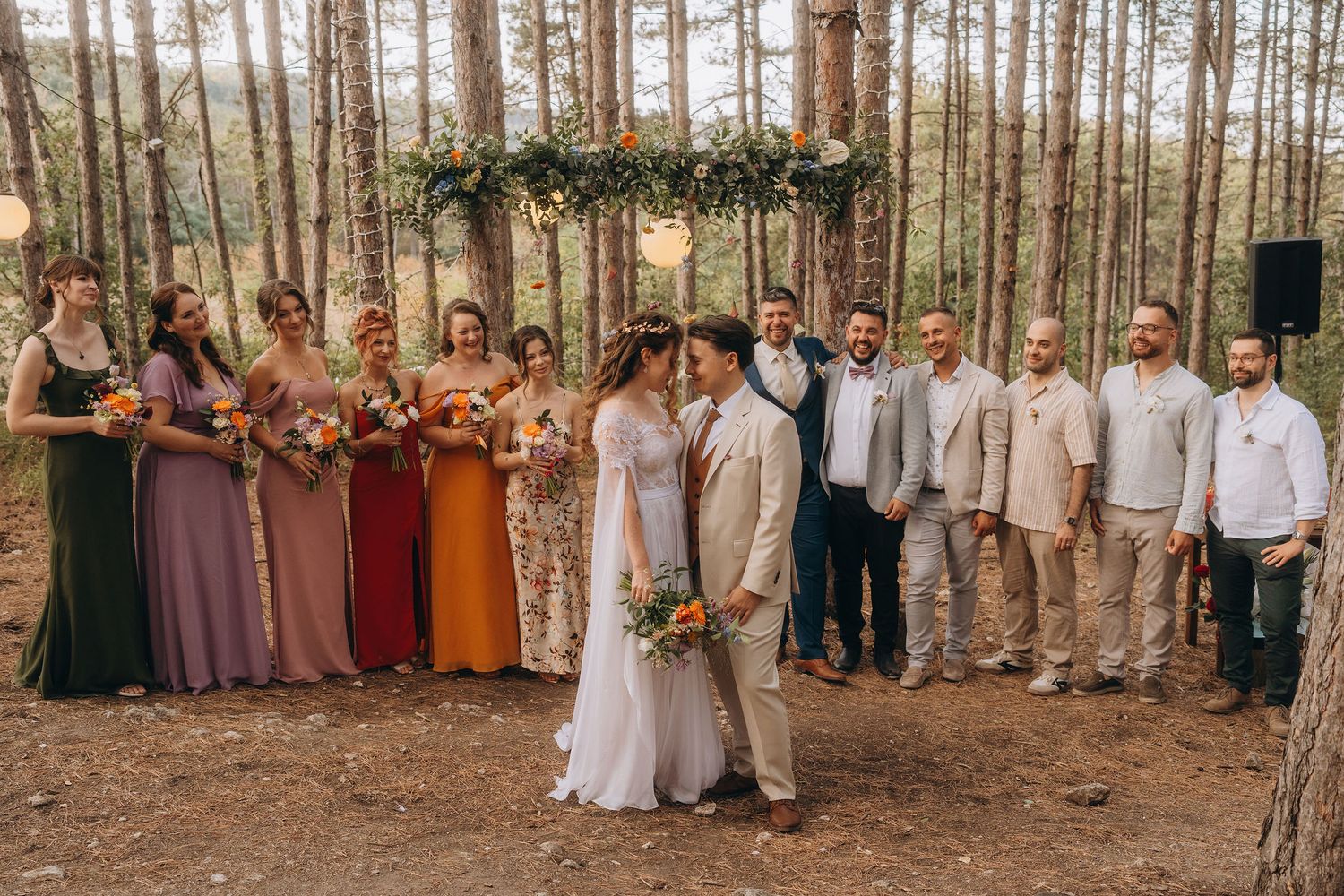 A bride and groom share a kiss surrounded by bridesmaids and groomsmen in a pine forest ceremony.