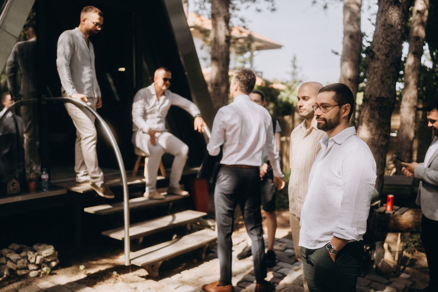 A group of well-dressed men in white shirts and gray trousers socializing outdoors near a modern A-frame structure.