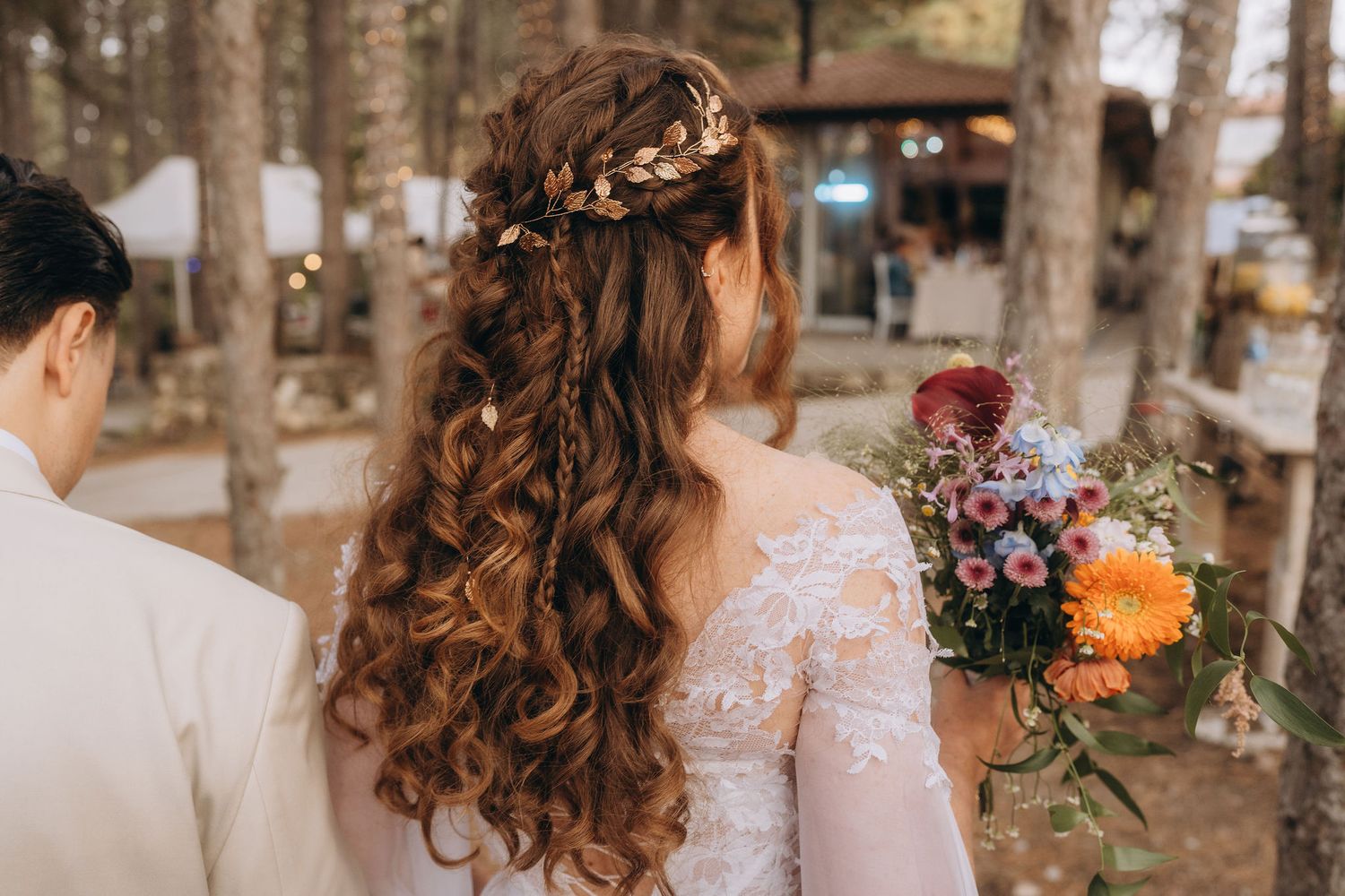 Back view of bride with curly hair, floral crown, lace dress, holding colorful bouquet in forest.