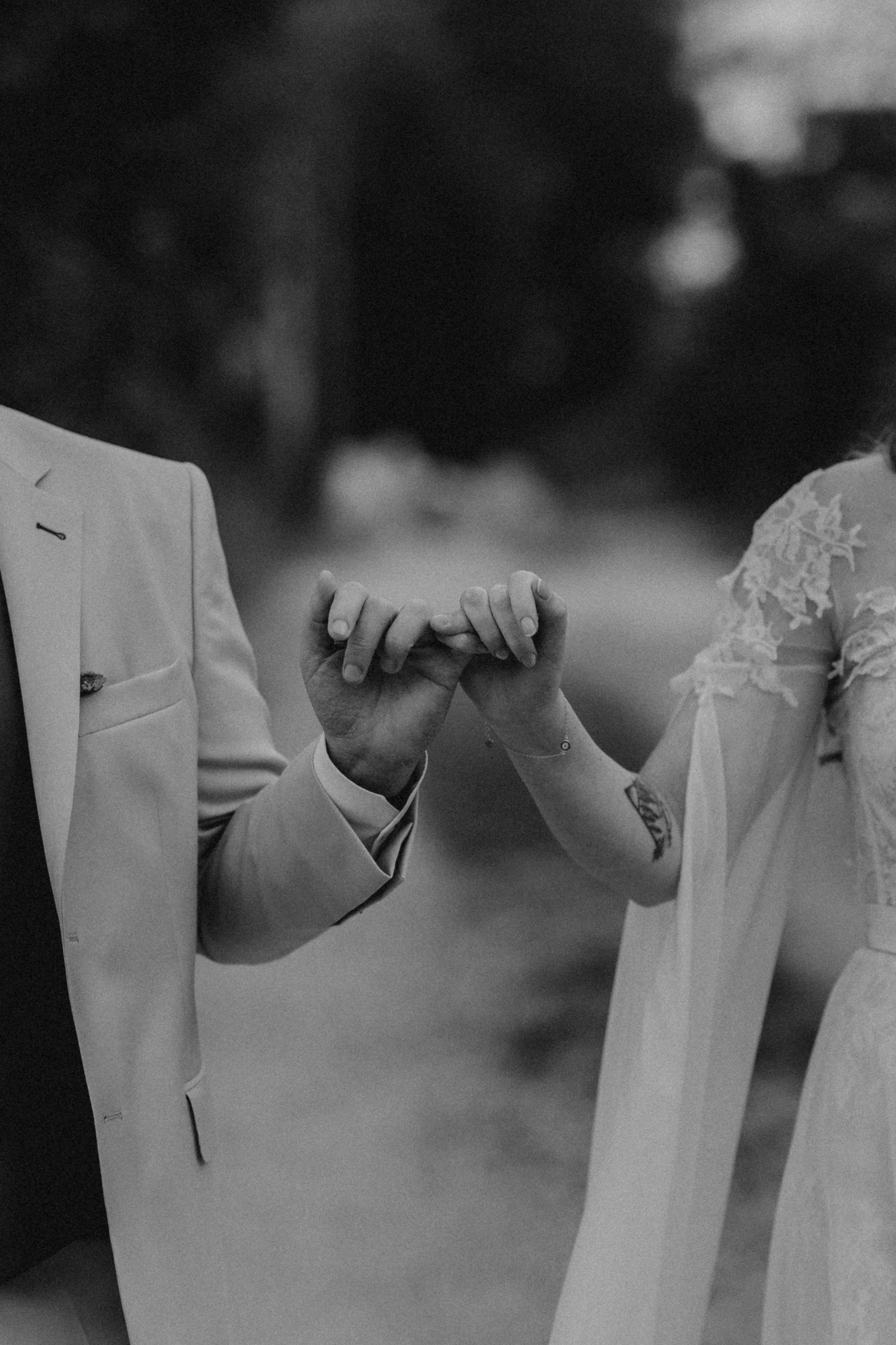 Black and white close-up of bride and groom holding hands, showing tattoo and lace wedding dress detail.