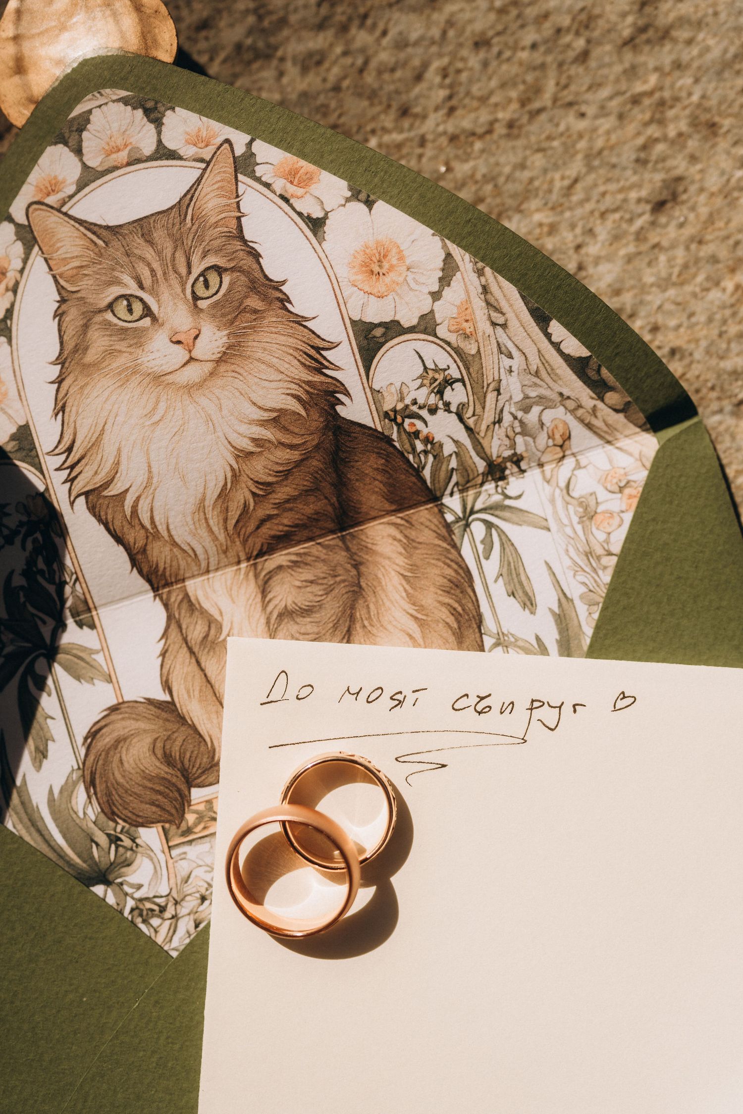Two gold wedding rings rest on a note beside a decorative cat illustration envelope in warm natural light.