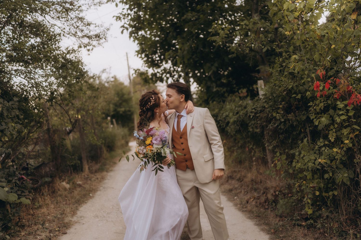 Bride and groom sharing a kiss on a garden path surrounded by lush green trees.
