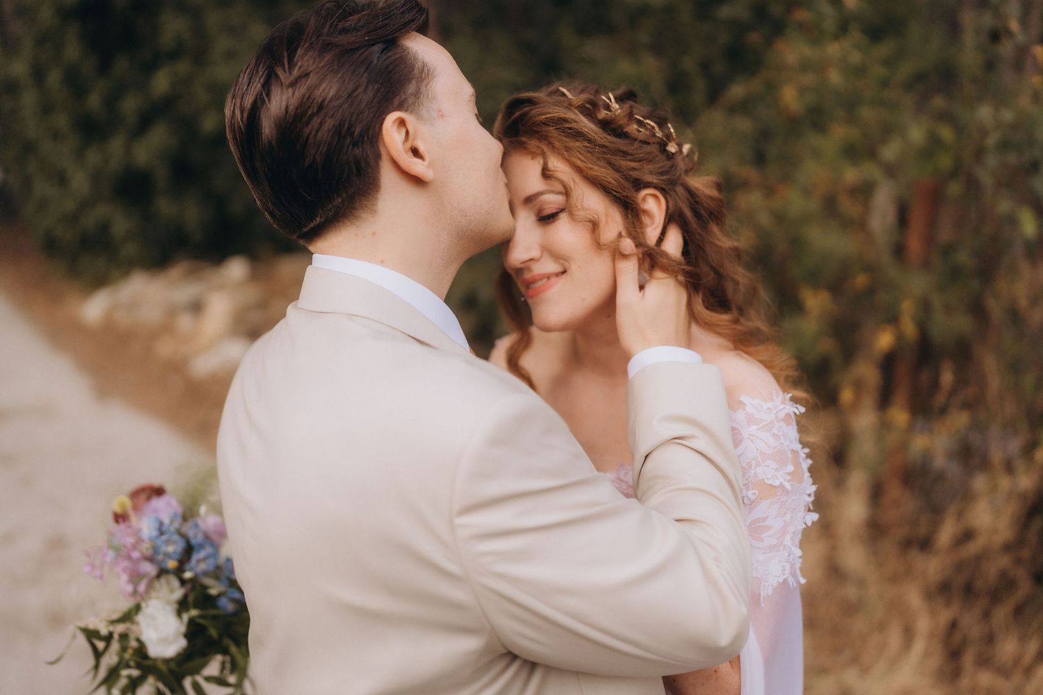 Groom gently kissing smiling bride's temple during golden hour outdoor wedding portrait in nature.