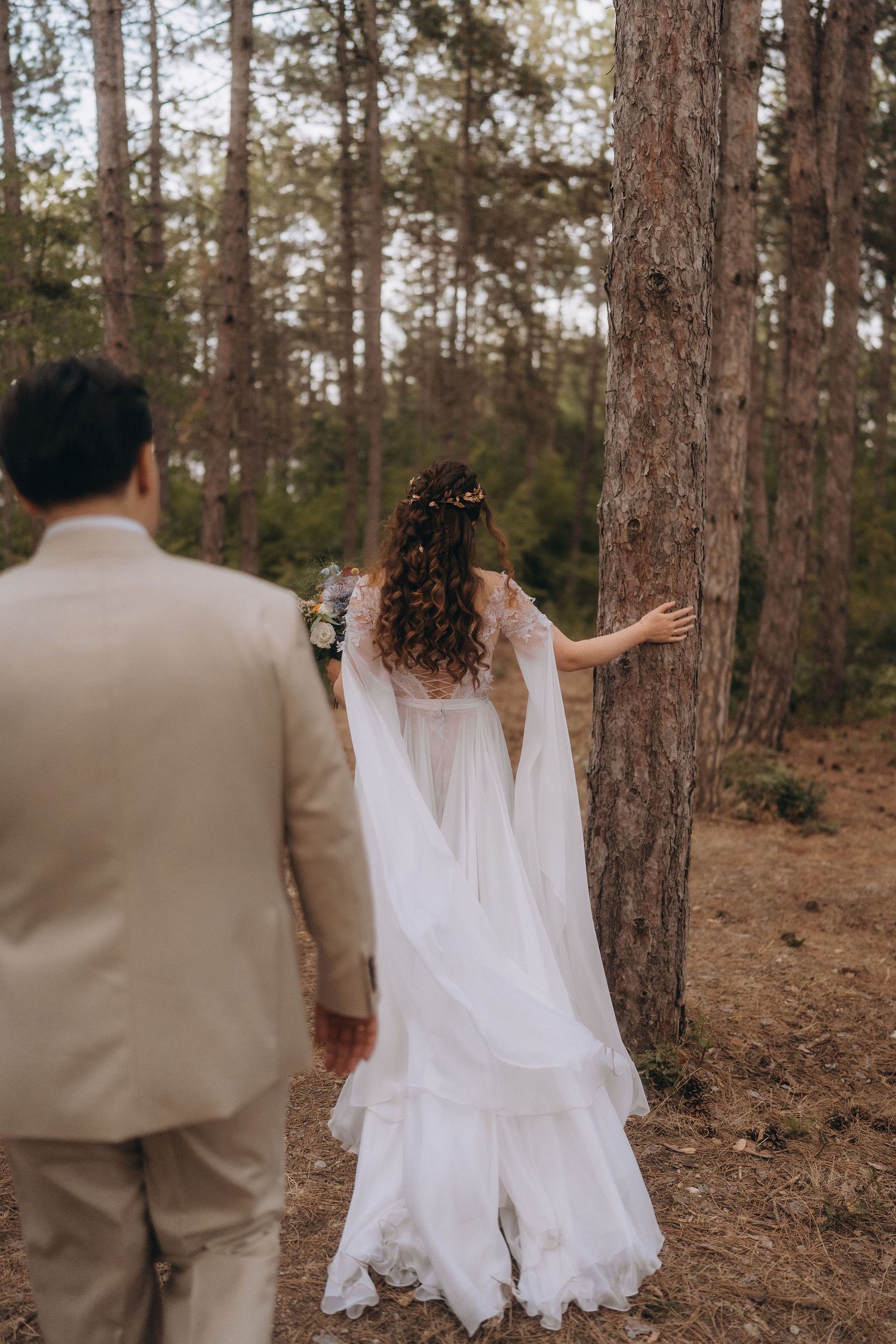 Bride in flowing white cape dress with floral crown leans against pine tree as groom watches in forest setting.