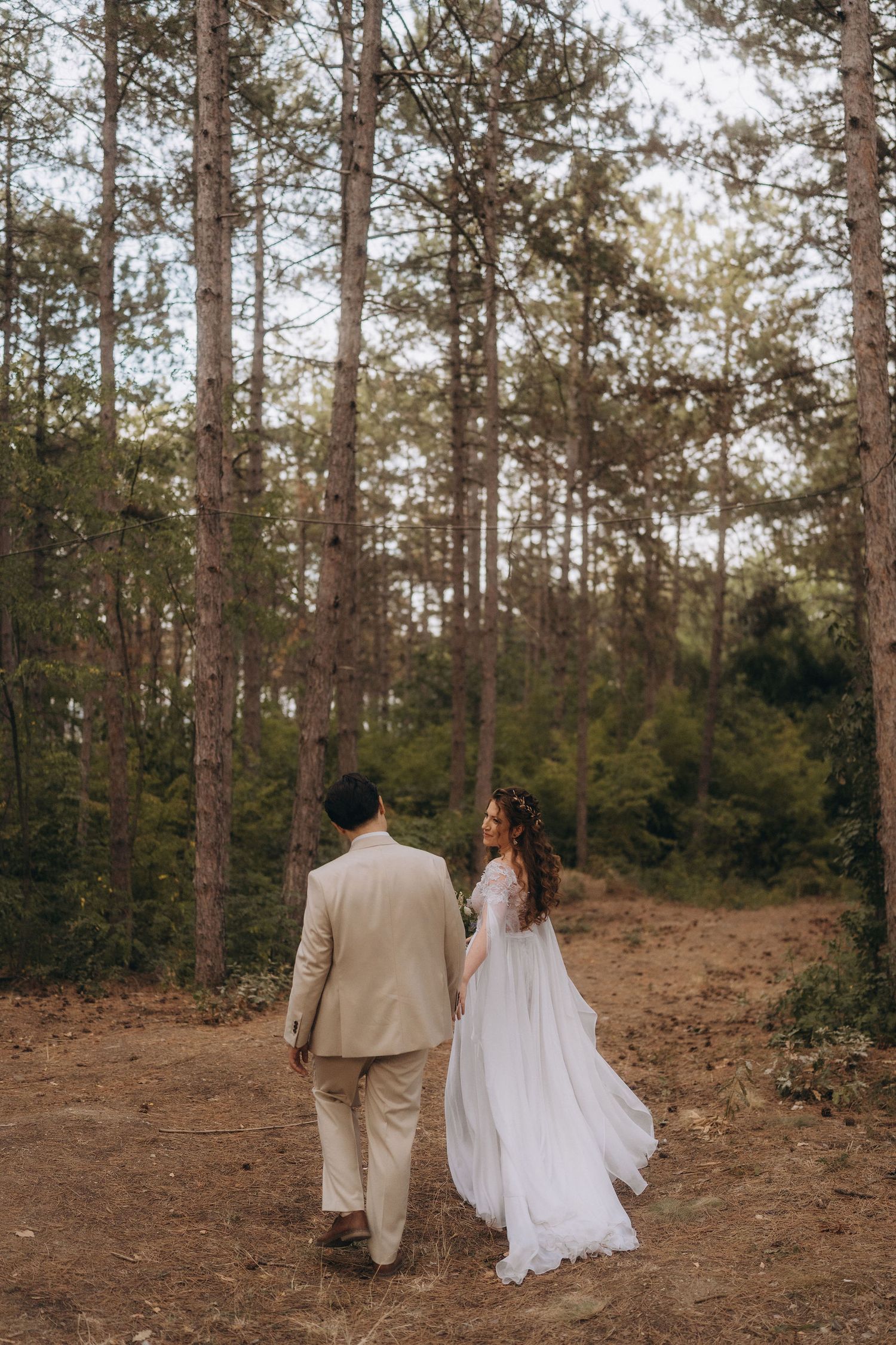 Bride in white gown and groom in beige suit walk hand in hand through pine forest.