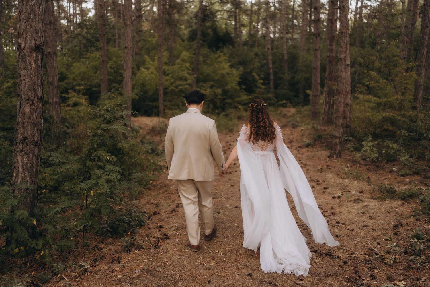 Bride and groom walking hand in hand through a lush pine forest, wedding veil flowing behind.