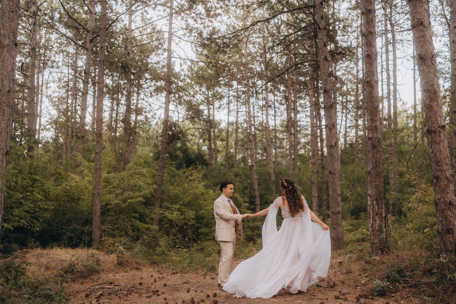 Groom twirls bride in flowing white dress among tall pine trees in forest wedding.