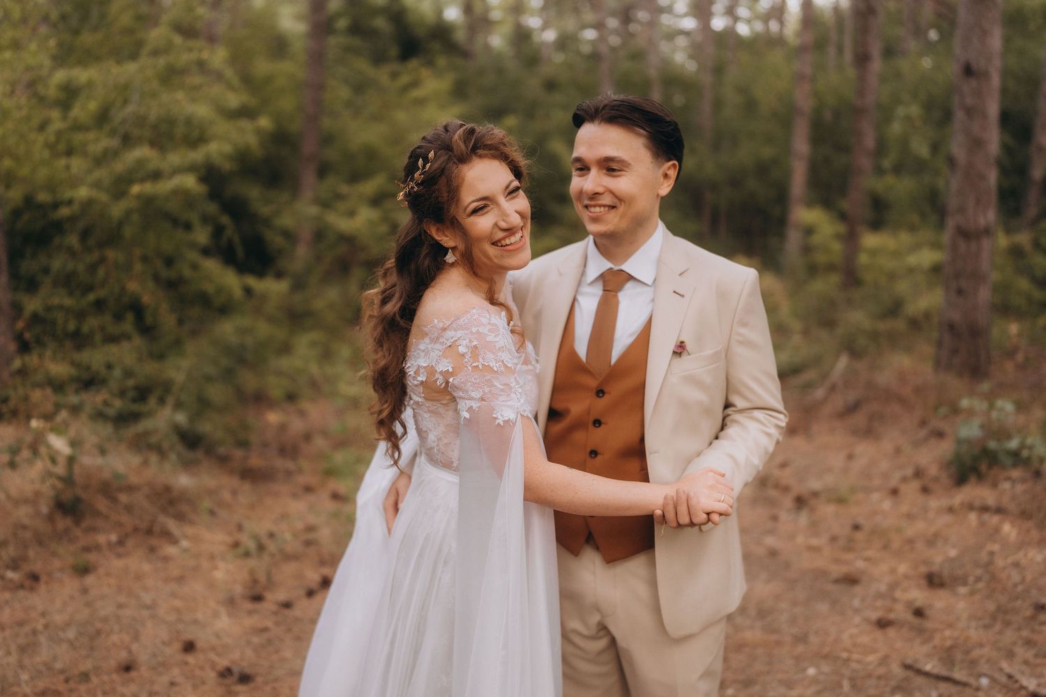 Smiling bride and groom embrace closely in a lush green pine forest wedding portrait.