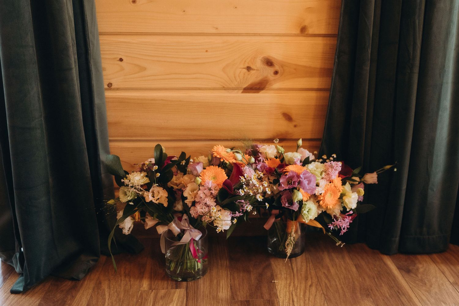 Three colorful wildflower bouquets in glass jars arranged on a wood floor against a pine wall with dark curtains.