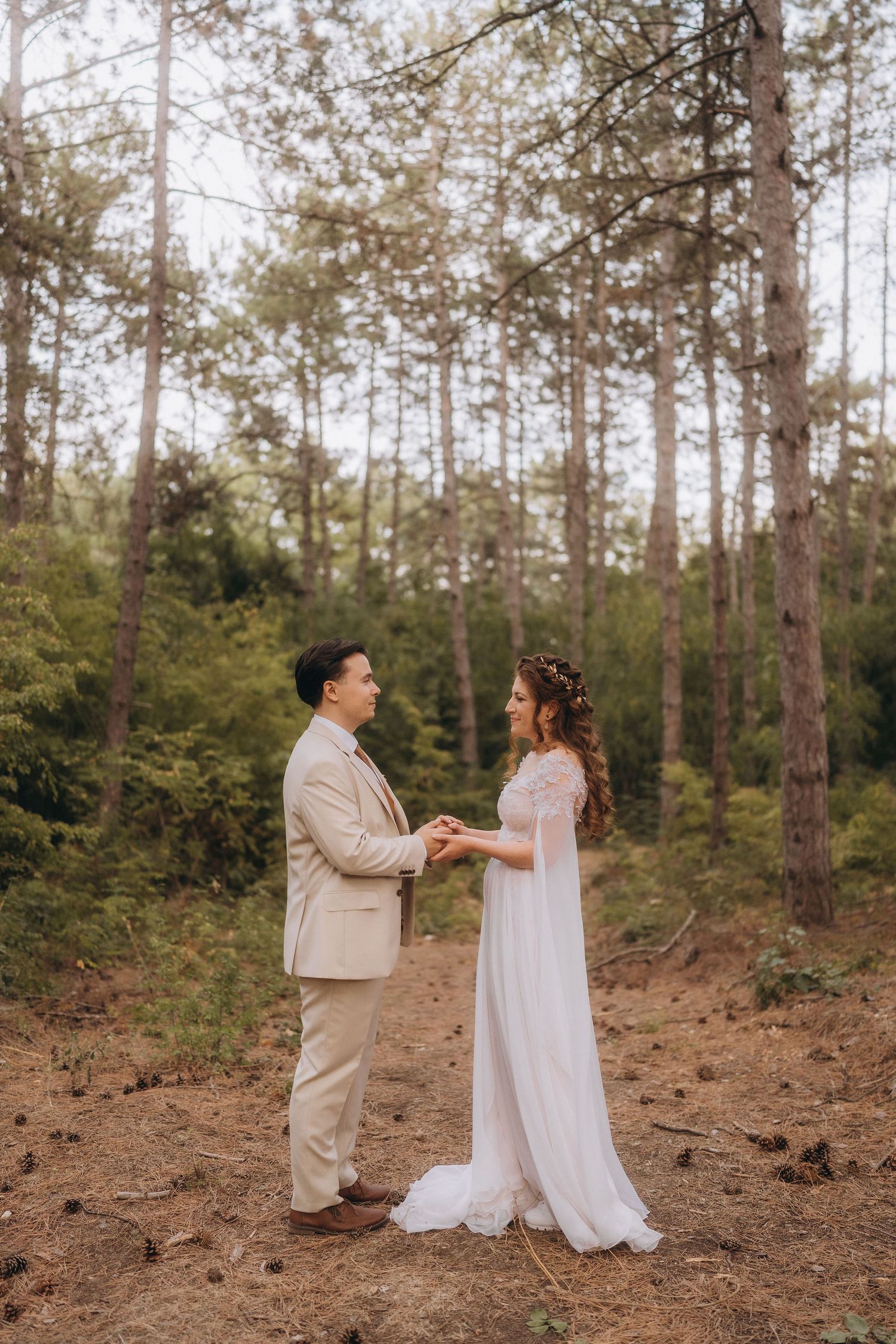 Bride and groom hold hands facing each other in a romantic forest wedding ceremony among tall pine trees.