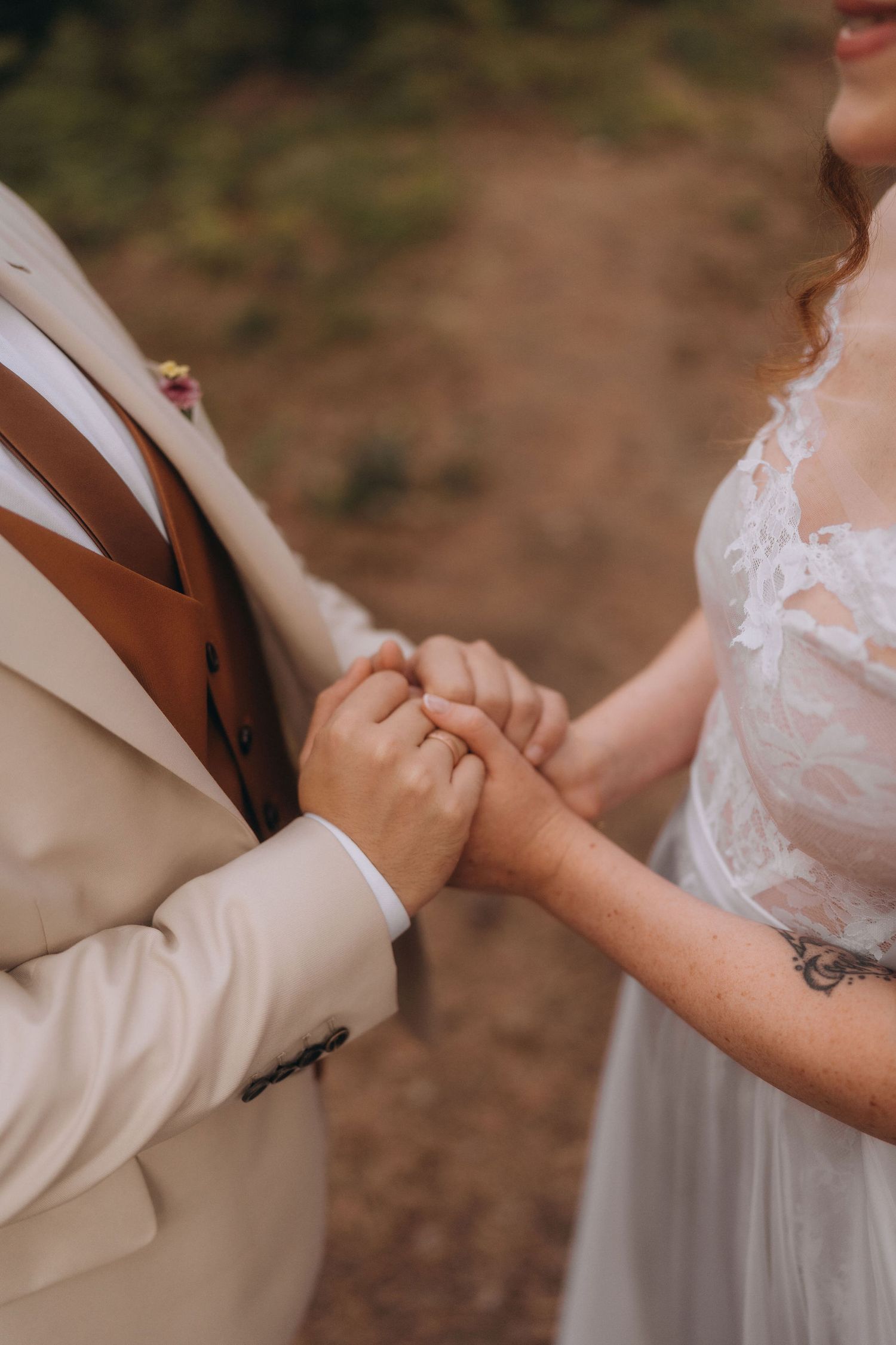 Bride and groom holding hands during wedding ceremony, groom in beige suit with brown vest, bride in lace white dress.