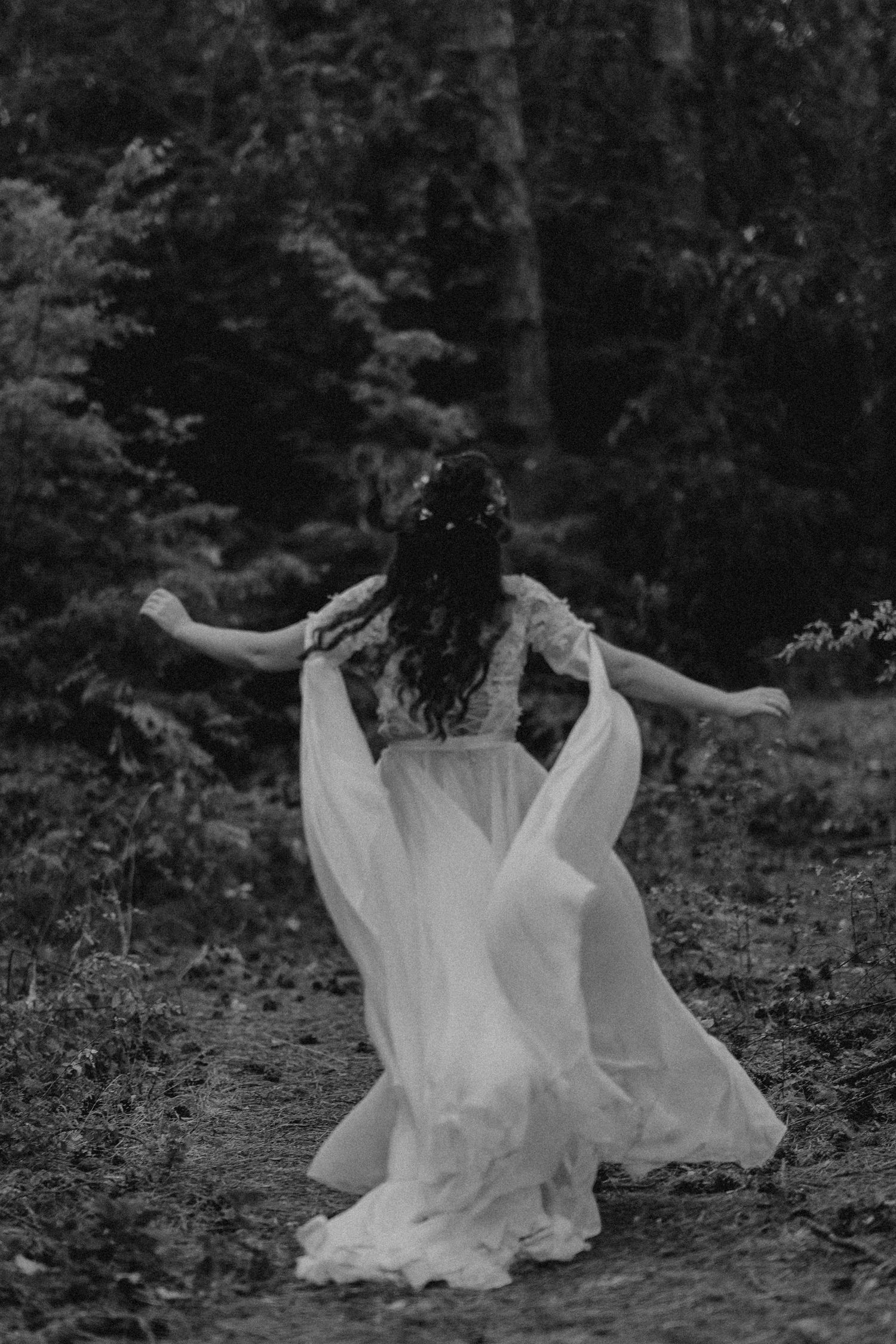 Black and white photo of bride in flowing white gown with floral crown walking through forest path.