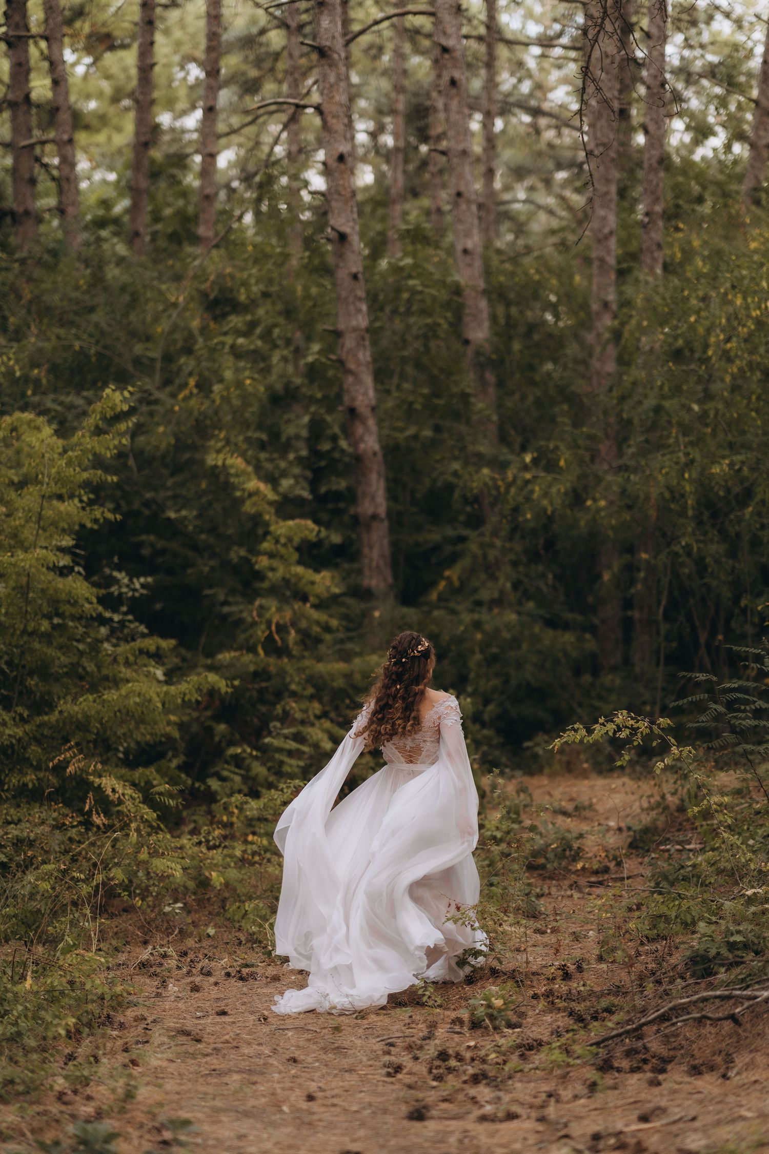 A bride in a flowing white wedding dress walks through a lush green forest path, her gown billowing in the breeze.