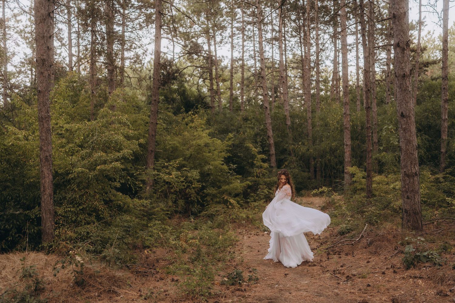 Bride in flowing white gown twirling in a lush forest setting with tall pine trees and green foliage.
