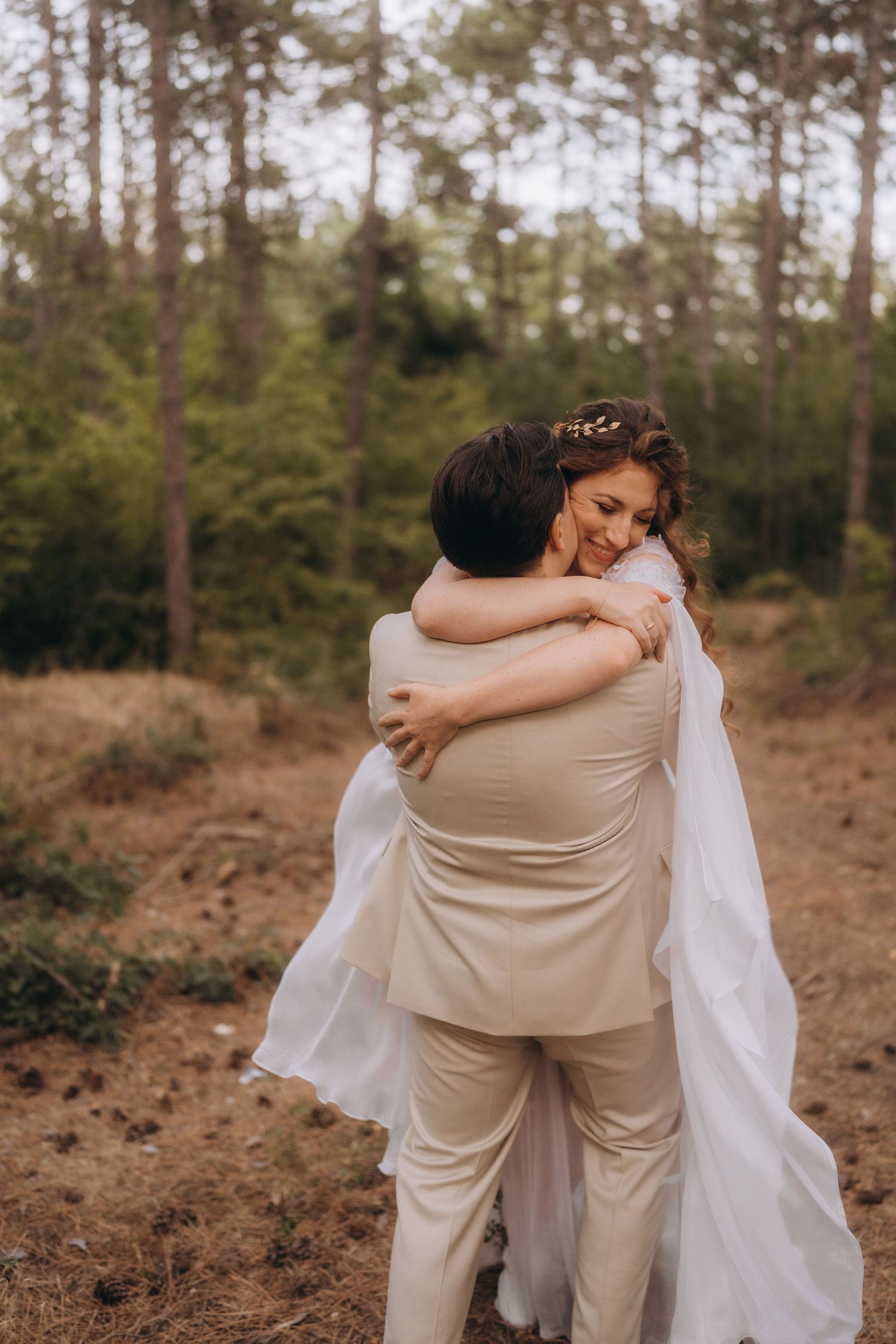 Bride and groom share a romantic embrace in a forest setting, with the bride in a flowing white cape dress.