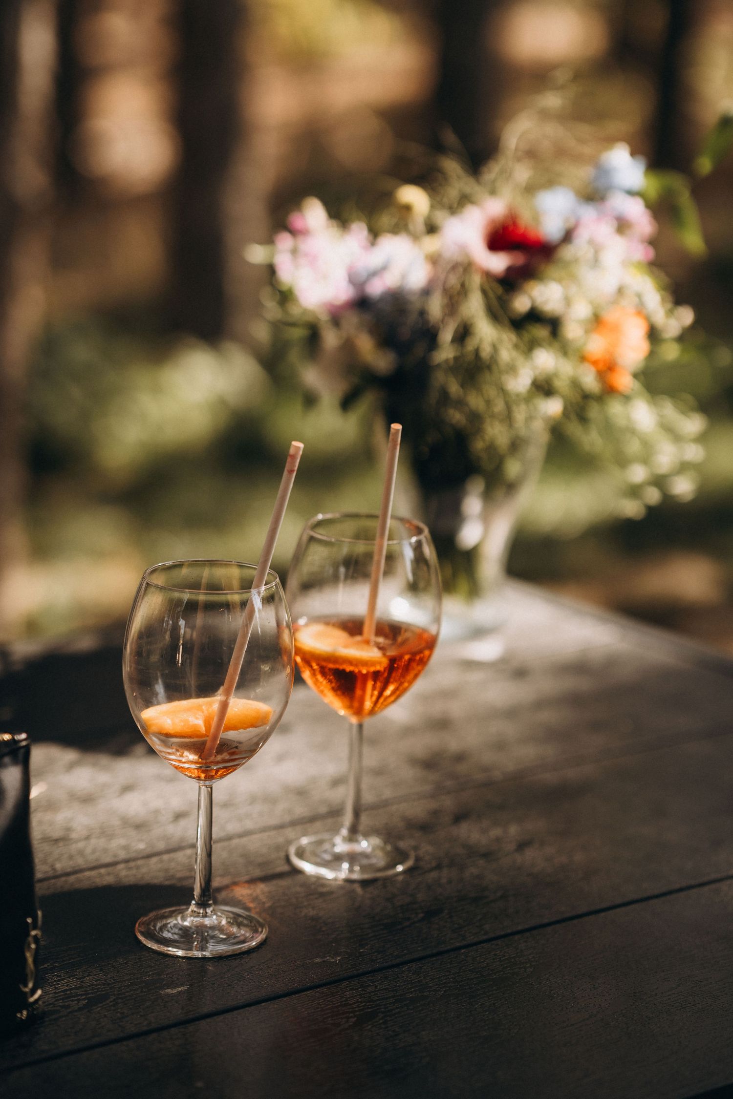 Two elegant wine glasses with orange aperol spritz cocktails and straws on a dark wooden table with a floral bouquet backdrop.