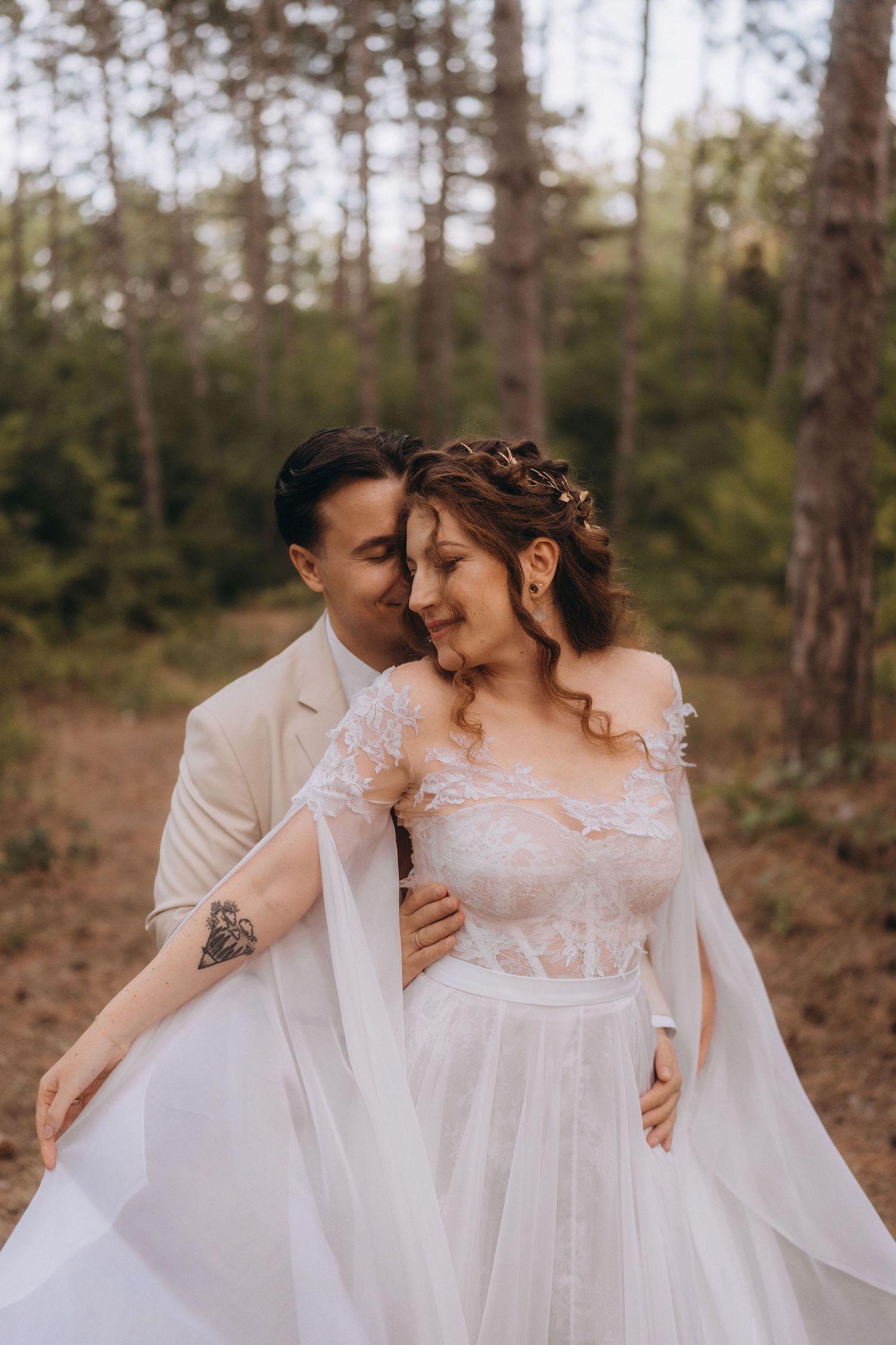 Groom in beige suit embraces bride in lace wedding dress tenderly in a sunlit pine forest setting.