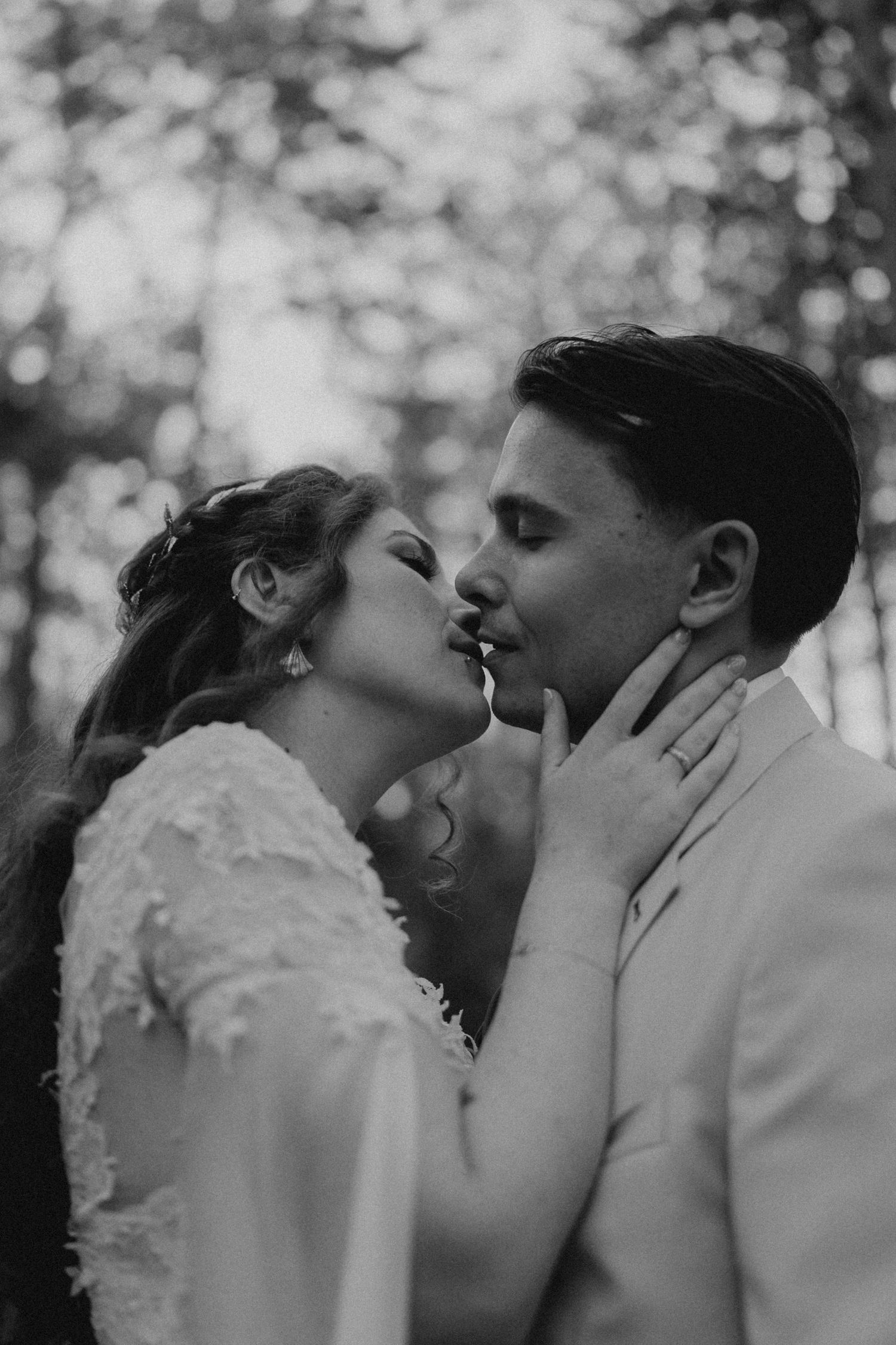 Black and white photo of a bride and groom sharing a romantic kiss outdoors among trees on their wedding day.