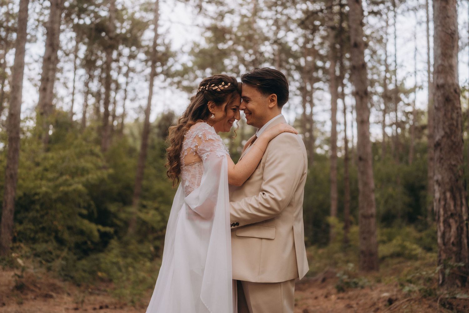 A bride and groom share a romantic moment in a forest, dressed in a white gown and beige suit.